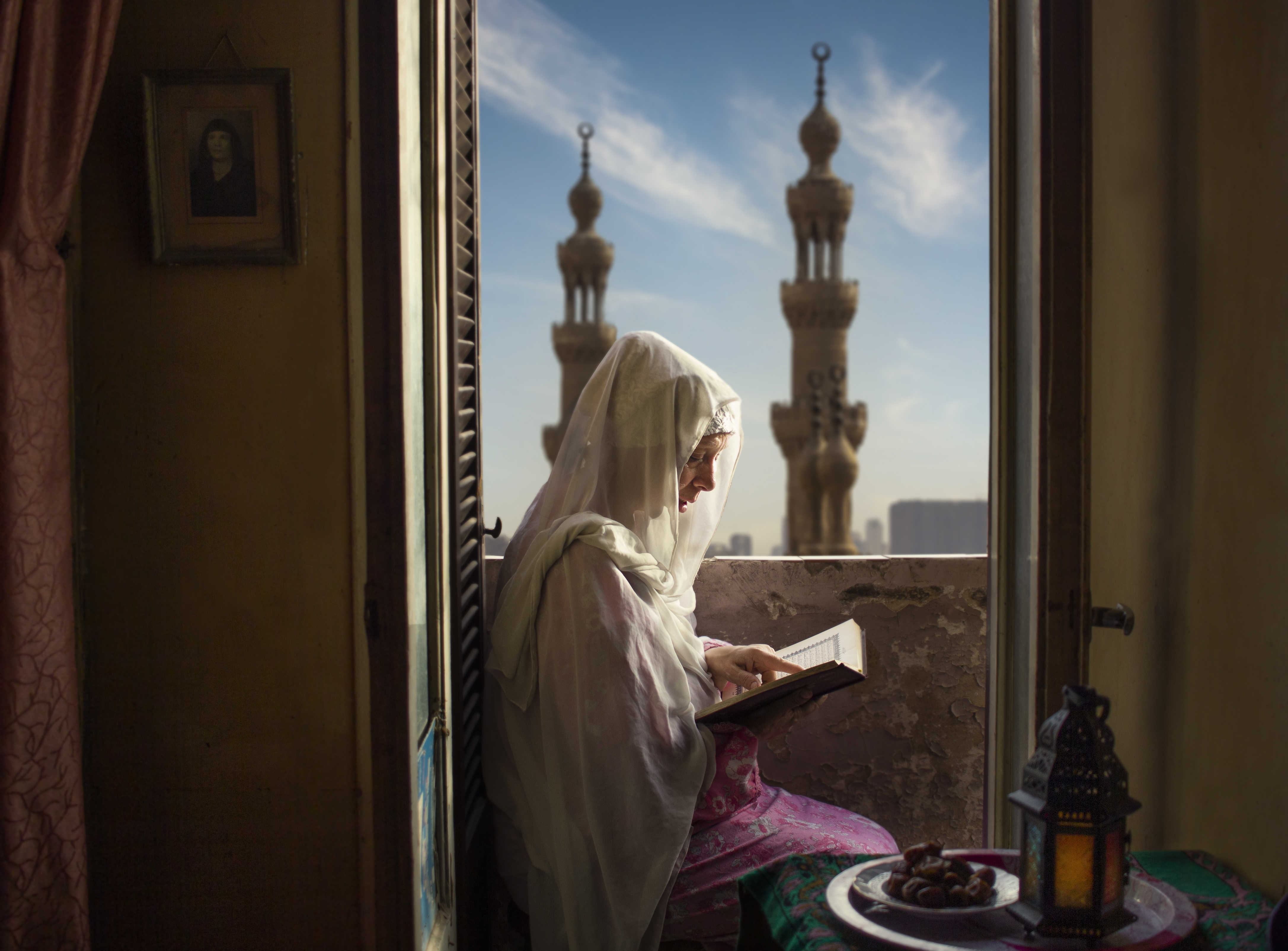A woman in Egypt is inside her house practicing her rituals of worship during the month of Ramadan. The two taller minarets appear to be part of Al-Rifa'i Mosque in Cairo.