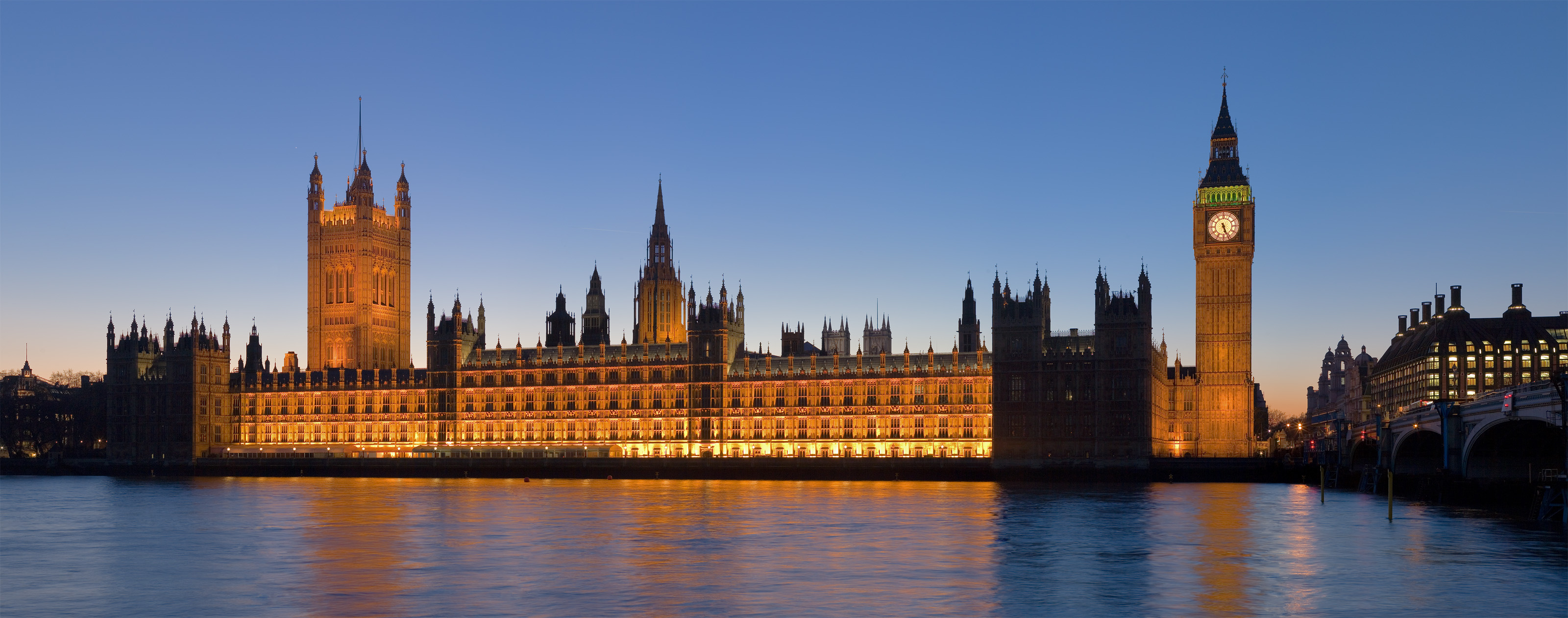The Palace of Westminster at night, as seen from the opposite side of the River Thames. Victoria Tower and the House of Lords are on the left, while the Clock Tower of Big Ben and the House of Commons are on the right. The spire left of center is the 300-foot ventilation chimney above the central lobby. The twin towers, with a flagless pole just visible in the background, belong to Westminster Abbey.